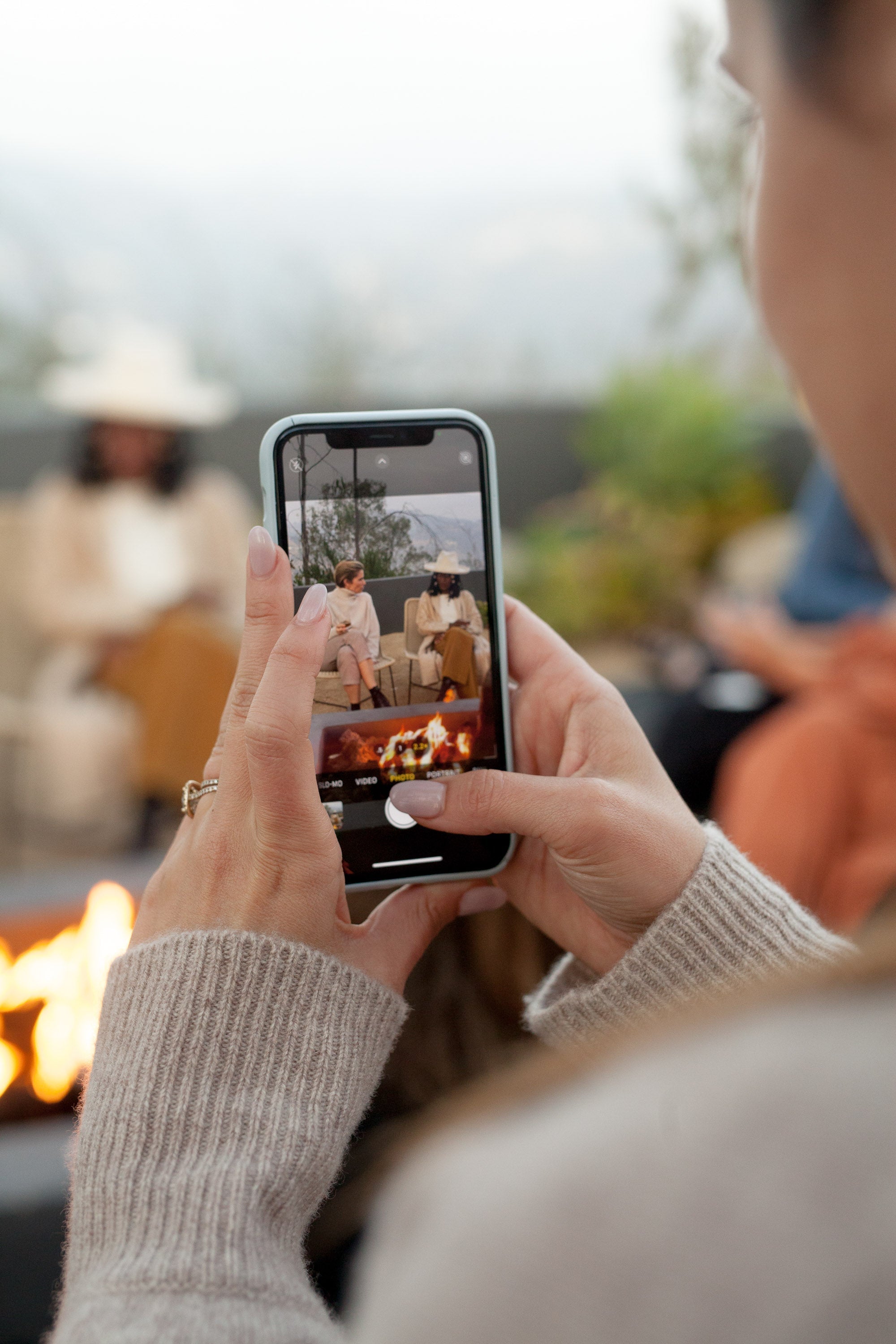 Person taking a photo with their phone of a bonfire gathering outdoors, wearing a cozy sweater