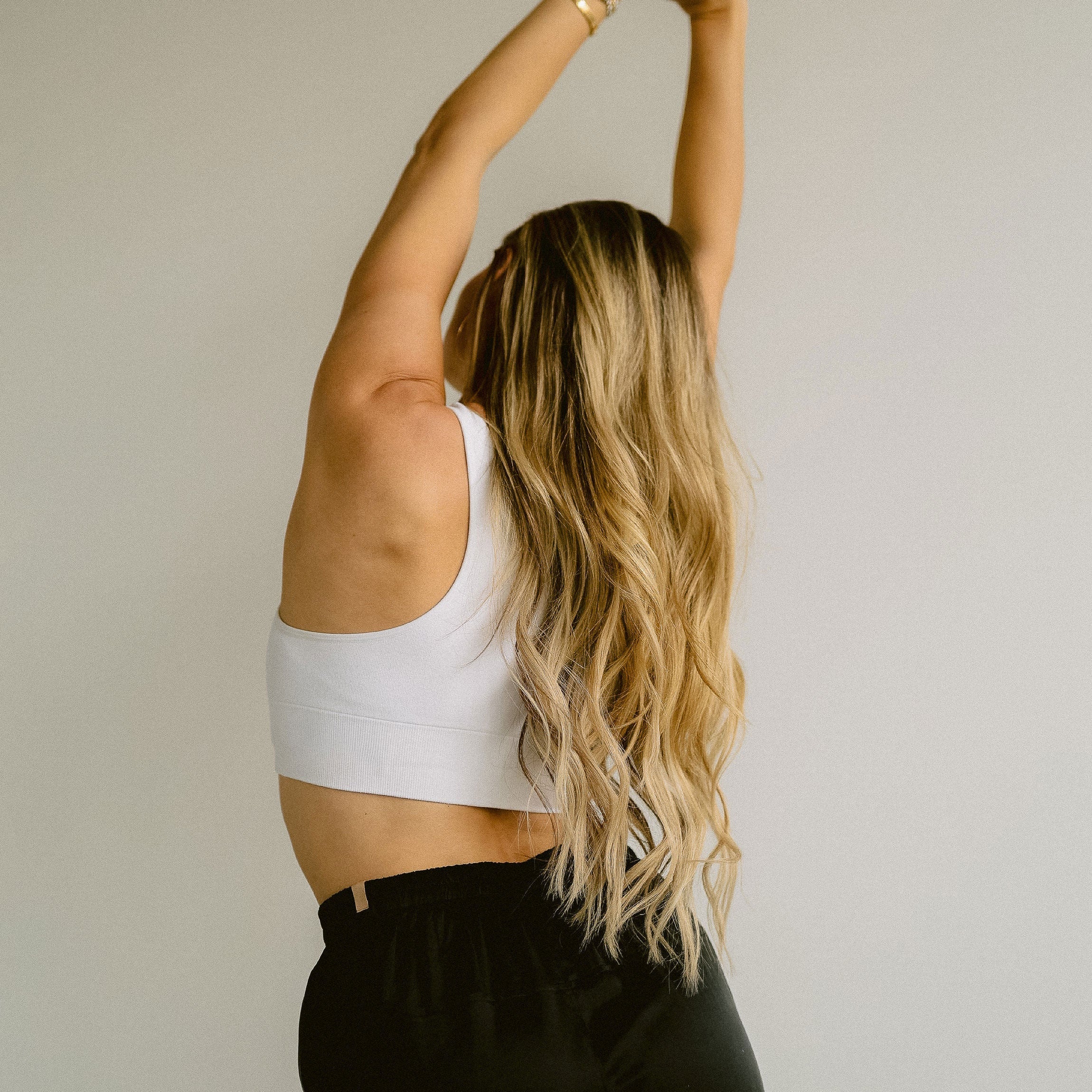 Woman with long blonde hair stretching with arms raised overhead, wearing a white crop top