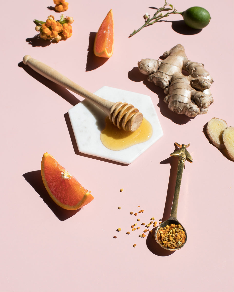 Honey dipper, ginger root, orange wedges and a spoon of bee pollen on a pink surface