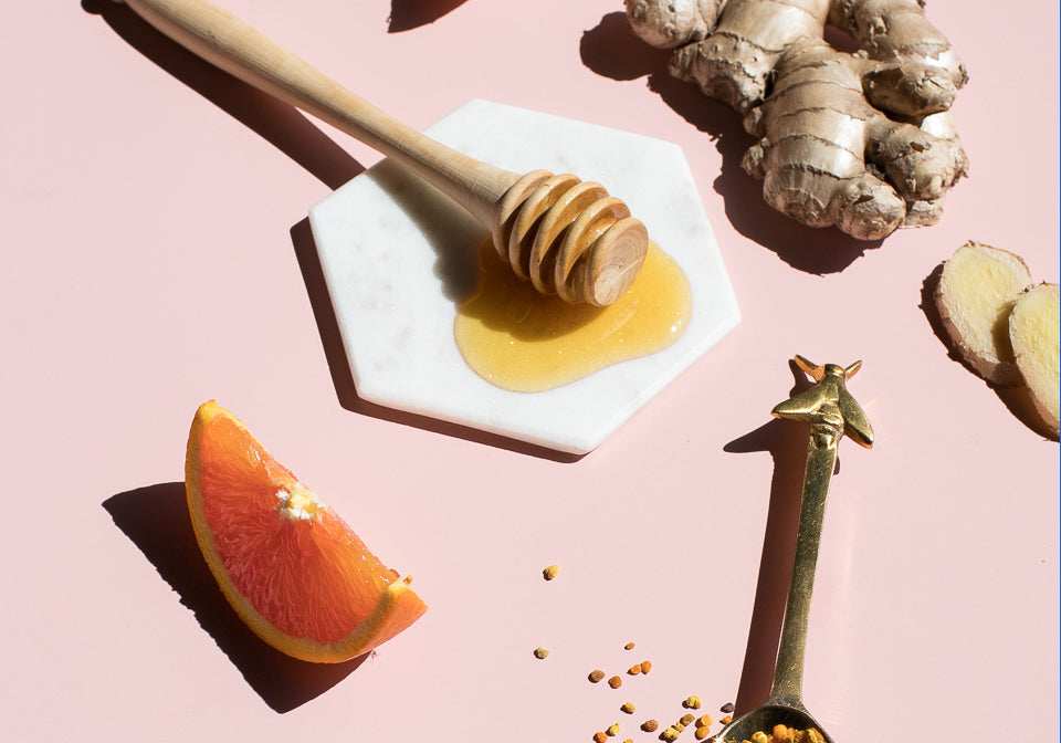 Honey dipper, ginger root, orange wedges and a spoon of bee pollen on a pink surface