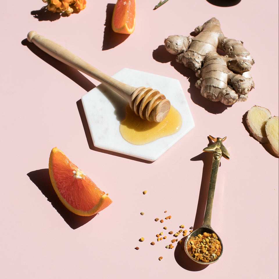 Honey dipper, ginger root, orange wedges and a spoon of bee pollen on a pink surface
