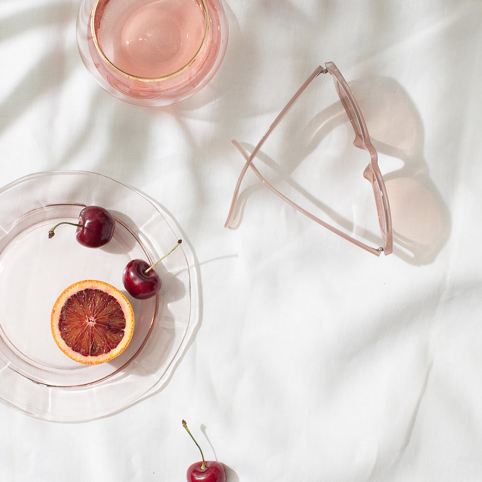 Cherries and a blood orange half on a glass plate beside a pink drink and sunglasses