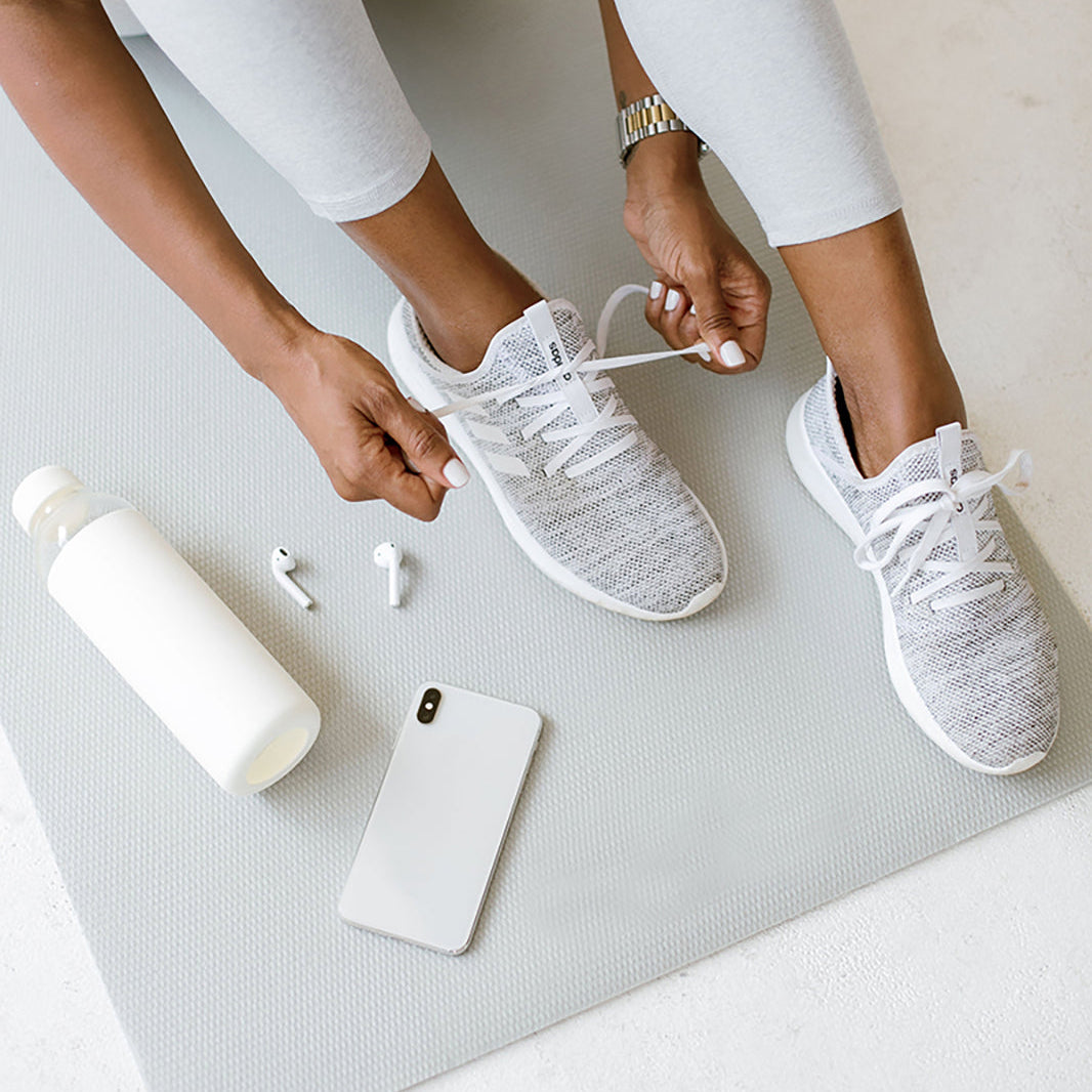 Overhead view of a woman tying her running shoes on a yoga mat, with a water bottle and phone beside her