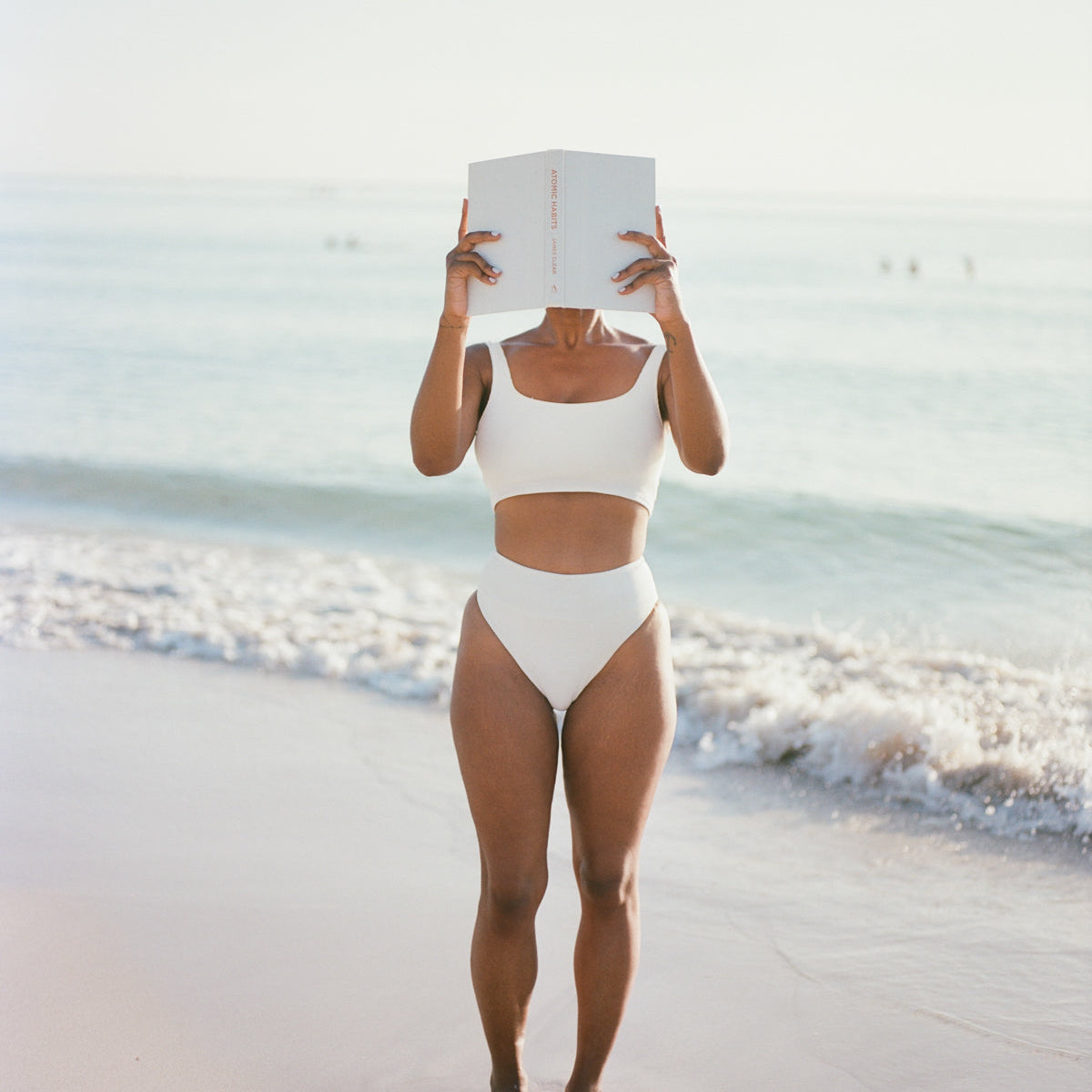 Woman standing on a beach shoreline in a white swimsuit, holding an open book up over her face