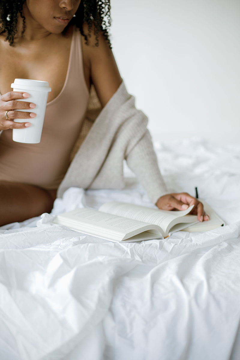 Woman sitting on a white bed with a coffee, writing in a journal