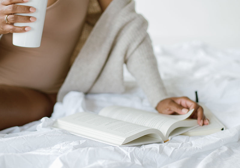 Woman sitting on a white bed with a coffee, writing in a journal
