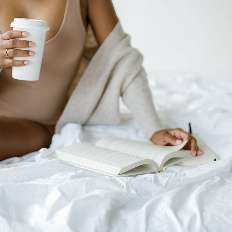 Woman sitting on a white bed with a coffee, writing in a journal