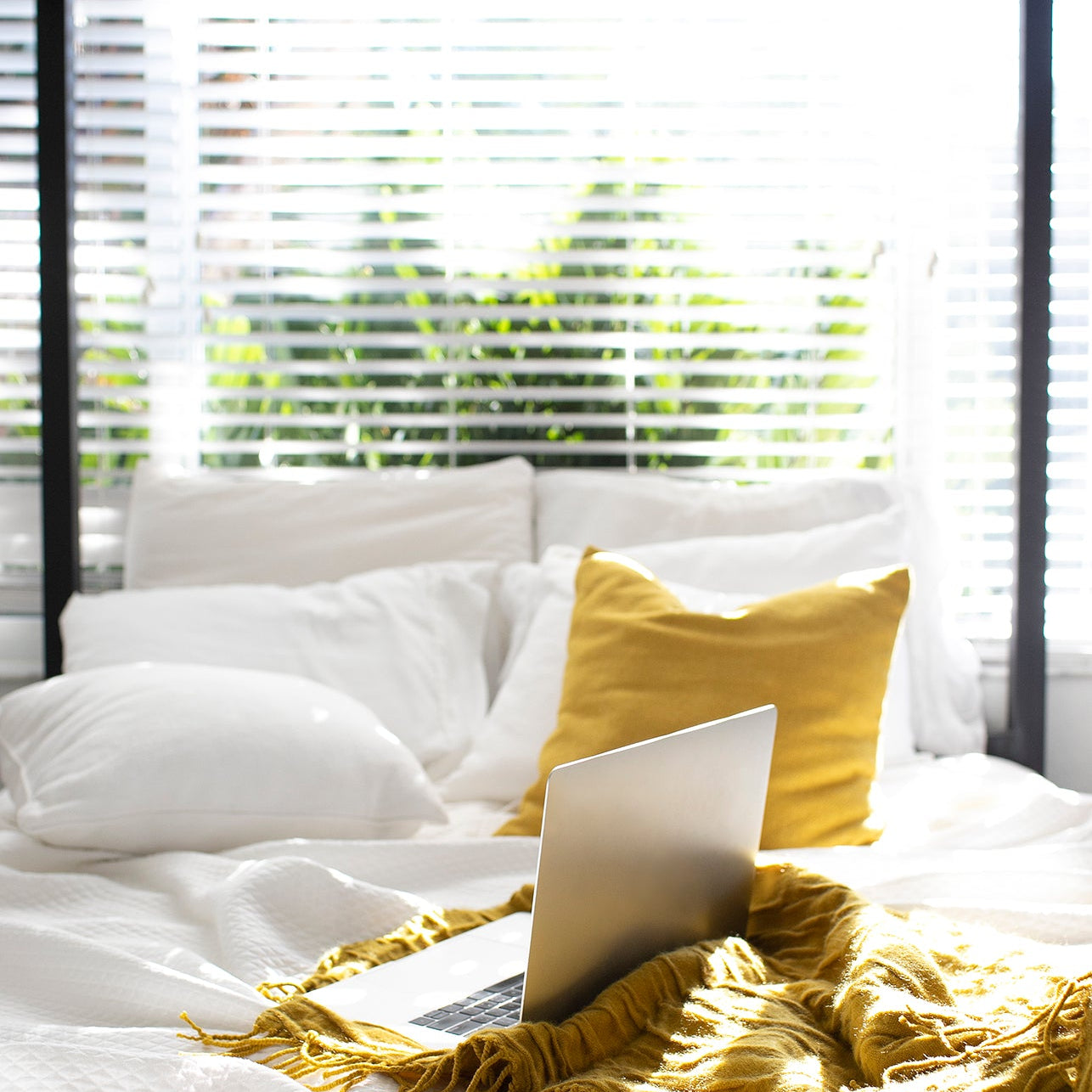 Sunlit bedroom with white bedding, a yellow throw and a laptop on the bed