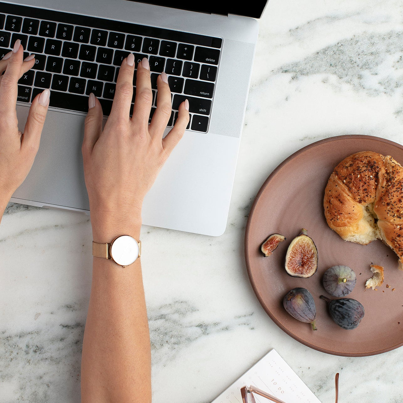 Hands typing on a laptop beside a plate of figs and bread