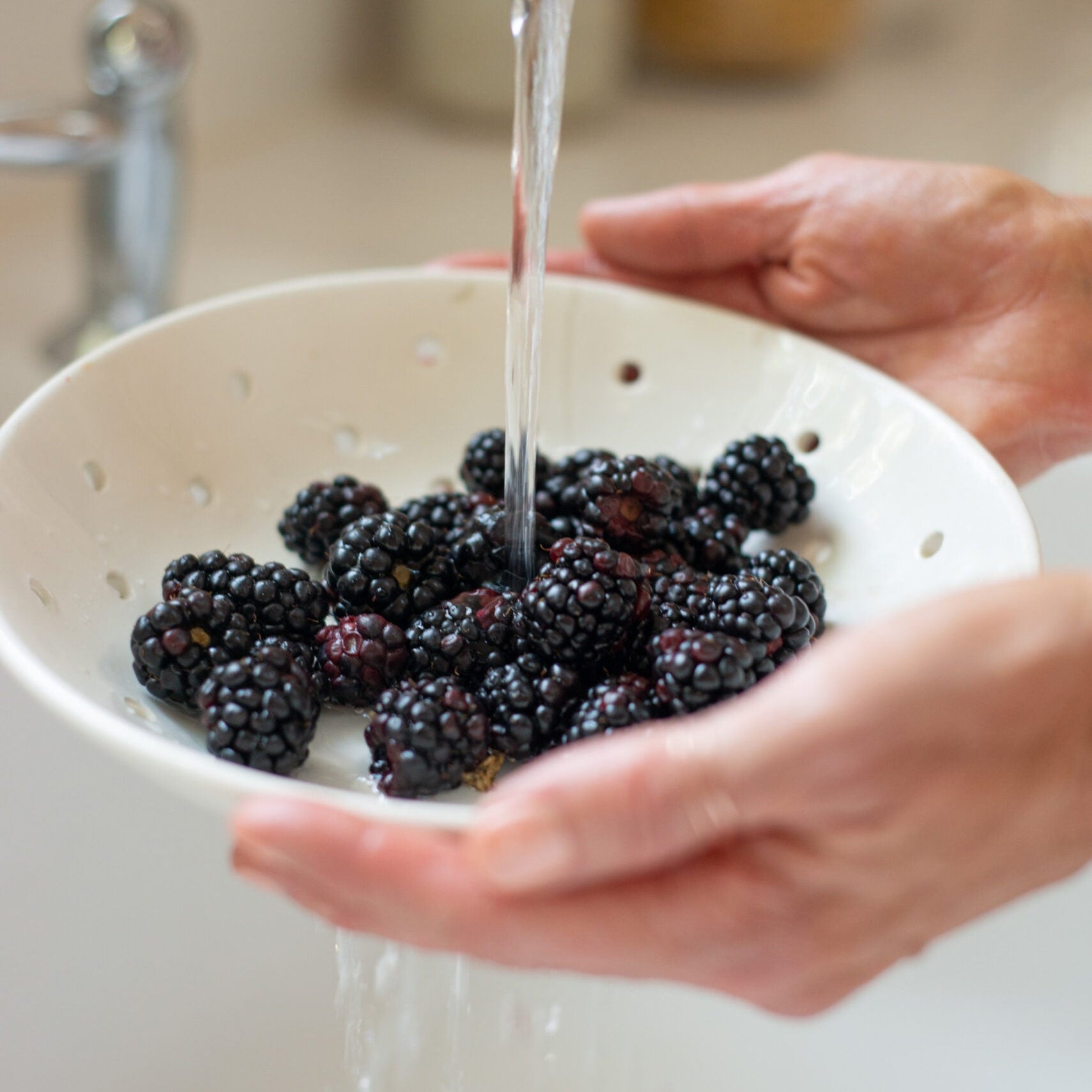 Hands rinsing blackberries in a colander under a tap