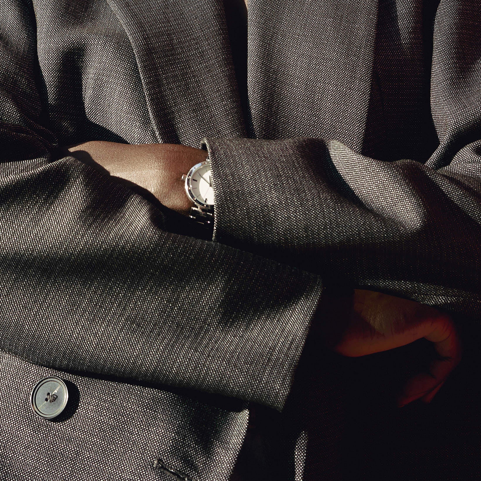 Close-up of a person in a tailored gray blazer with arms crossed, wearing a silver wristwatch