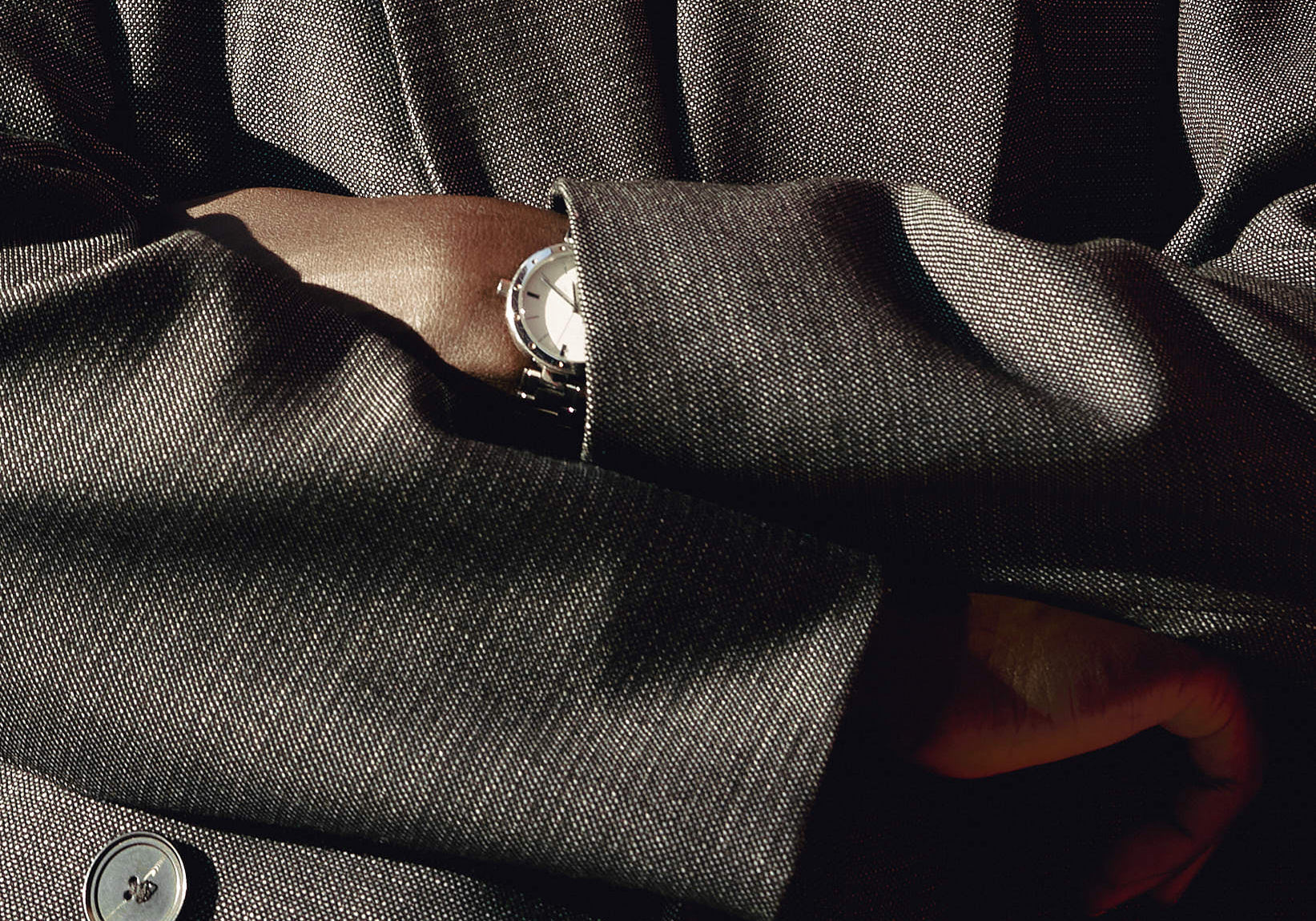 Close-up of a person in a tailored gray blazer with arms crossed, wearing a silver wristwatch