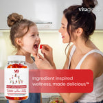 A woman feeding a young girl a red gummy vitamin, with a bottle of VG Kids Multivitamins in the foreground and a red text banner reading 'Ingredient inspired wellness, made delicious'.