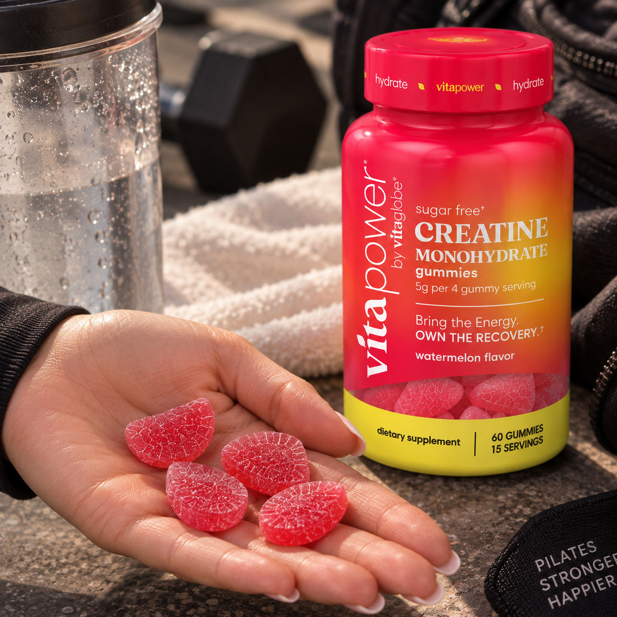 Hand holding 4 wedge-shaped VitaPower Sugar-Free Creatine Gummies in Watermelon flavor next to the bottle, with a water glass, dumbbell and towel on a gym floor