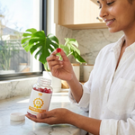 Woman holding a supplement bottle and a red capsule in a kitchen setting