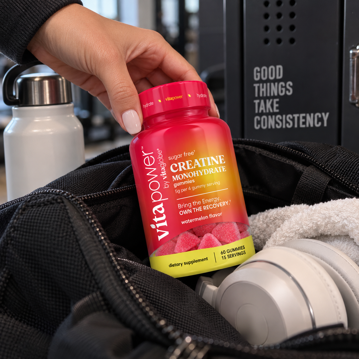Hand pulling a bottle of VitaPower Sugar-Free Creatine Monohydrate Gummies, Watermelon flavor, out of a black gym bag with a towel, headphones and water bottle in a locker room